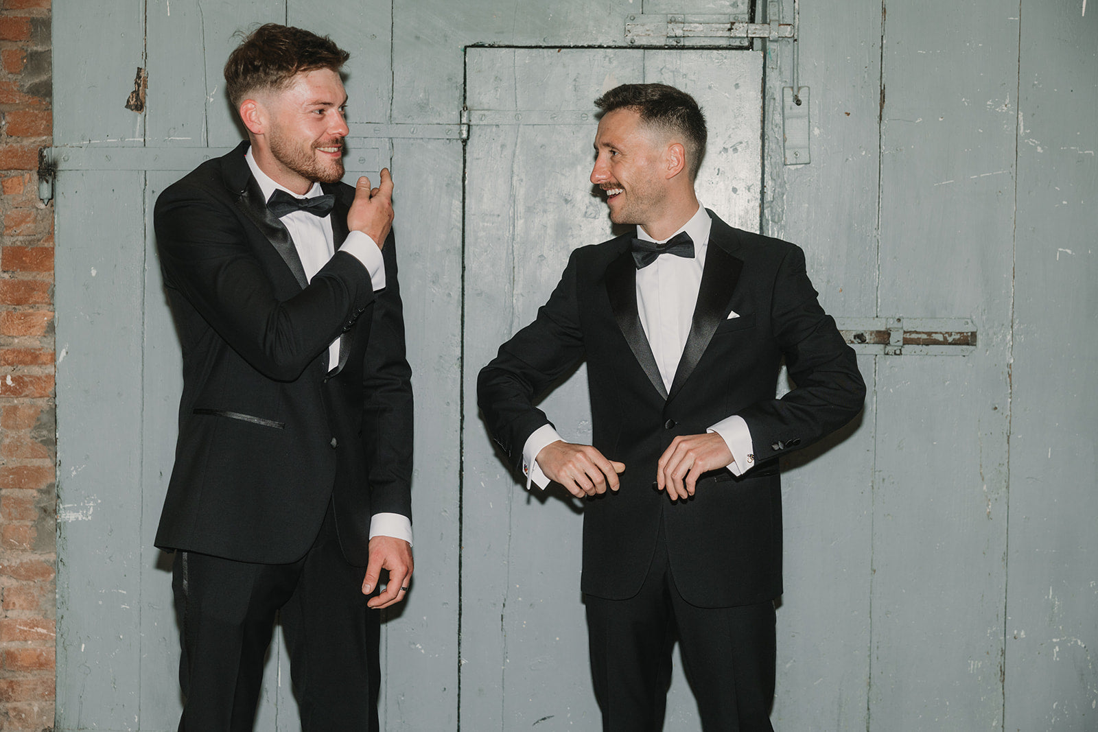 Two men in black tuxedos standing against a gray wall.